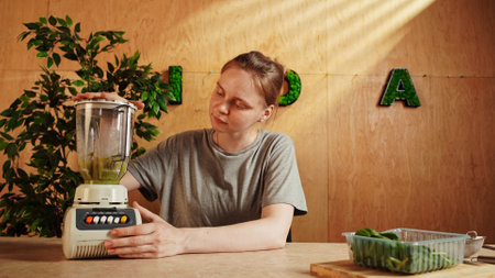 Woman sitting at the table at home in the kitchen and preparing healthy omelet for breakfast, mixing eggs, green leaves and cheese in blender glass cup.の写真素材
