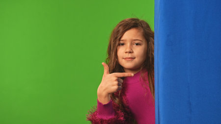 Little girl child peeks out and waves hand, pointing at blue space, positive expression, isolated on chroma key green screen background mockup.の写真素材
