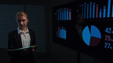 Young woman broker standing in office boardroom, analyzing financial data infographics on big display, holding folder with documents, checking information.の写真素材