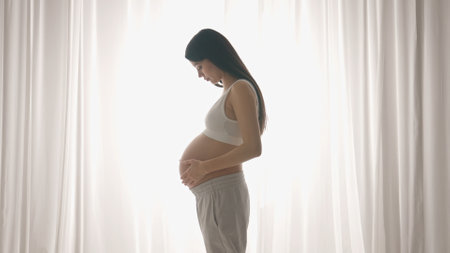Young happy Asian pregnant woman posing in studio with white curtains in background