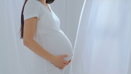 Young happy Asian pregnant woman posing in studio with white curtains in backgroundの写真素材