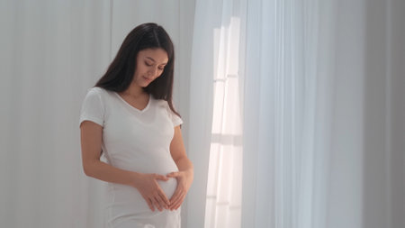 Young happy Asian pregnant woman posing in studio with white curtains in backgroundの写真素材