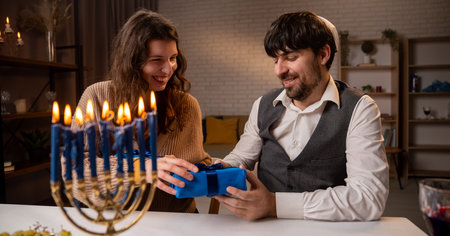 Jewish couple preparing for traditional hanukah celebrations, man and woman sitting at table, enjoying dinner and exchanging gifts, positive expression.の写真素材