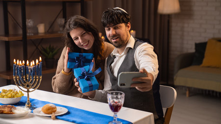 Jewish couple preparing for traditional hanukah celebrations, man and woman sitting at table, enjoying dinner, taking selfie with gifts on smartphone.の写真素材