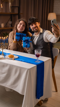 Jewish couple preparing for traditional hanukah celebrations, man and woman sitting at table, enjoying dinner, taking selfie with gifts on smartphone.の写真素材