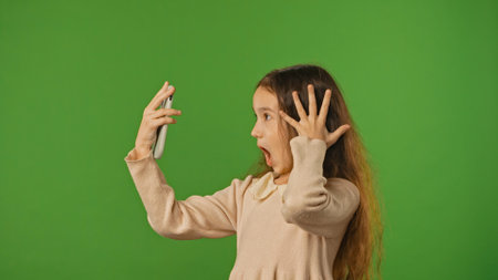 Little girl child holding smartphone and typing, scrolling social media, wow positive expression, isolated on chroma key green screen studio background mockup.の写真素材