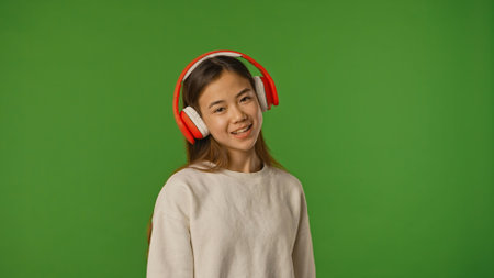 Asian girl child listening music in headphones, positive expression, isolated on chroma key green...
