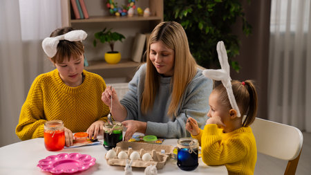 Mother and little daughters preparing for easter celebrations, sitting at table and making colored eggs with ink and water, smiling and having fun together.の写真素材