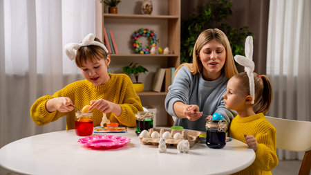 Mother and little daughters preparing for easter celebrations, sitting at table and making colored eggs with ink and water, smiling and having fun together.の写真素材