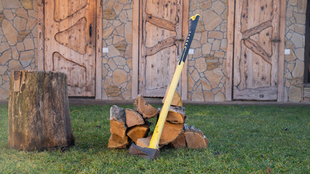 Shot of an axe leaning on pile of wood near the stump on green grass at rural house backyard at...