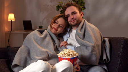 Lovely couple relaxing on couch at home, man and woman in the room watching tv, cuddling covered in plaid, talking and eating popcorn, positive happy expression.の写真素材