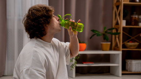 Young man in the morning in bedroom doing warmup, man sitting and drinking water from sport bottle, resting after physical exercises.の写真素材