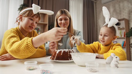Mother and little daughters preparing for easter celebrations, sitting at table and decorating festive cake, putting sugar glaze on top with spoons.の写真素材