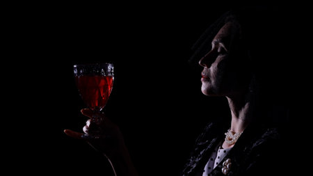Woman actress silhouette in retro outfit with pin-up hairstyle and makeup posing at camera isolated on black background, woman in veil holding glass of red wine.の写真素材