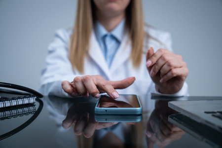Woman doctor in white coat sitting working at desk in the clinic office room, using smartphone, close shot of woman holding hand finger over smartphone screen.の写真素材