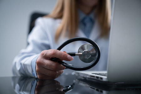 Woman doctor in white coat sitting working at desk in the clinic office room, working on laptop and holding stethoscope in hand, close up shot.の写真素材