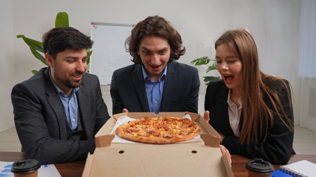 Business team of men and woman sitting at desk in the office having dinner break with food delivery, people with amazed happy expression looking at opened box with pizza.の写真素材