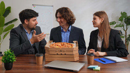 Business team of men and woman sitting at desk in the office having dinner break with food delivery, people talking and smiling, stack of pizza boxes standing on the table.の写真素材