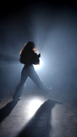 Silhouette of a woman dancing jazz-funk in a studio. Black background filled with smoke, white...