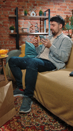 Bearded man spending time at home with fast food delivery, sits on couch eating chops and shopping online, typing card number on smartphone.の写真素材