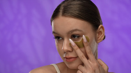 Young woman with makeup posing at camera in studio, smiling applying eye patch under eye, close up...