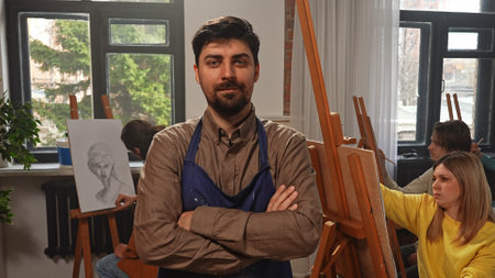 Man art teacher in apron posing in front of group of people drawing at easels during master class in...
