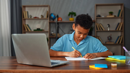 Young african american child boy studying at home, kid sits at desk attends school class online on laptop, listening lesson and writing information in notebook.の写真素材