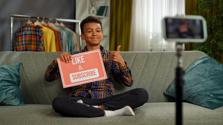 Young african american child boy spending time at home, kid blogger sits on sofa records video on...