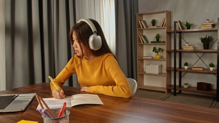 Focused student with headphones writing in notepad with laptop nearby