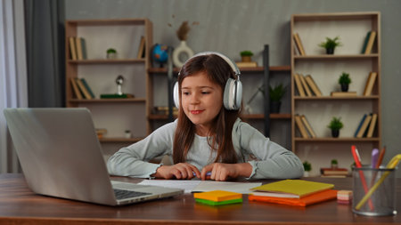 Young child girl studying at home, small girl sits at desk and attends school class online on laptop, doing homework, writing in notebook talking with teacher.の写真素材