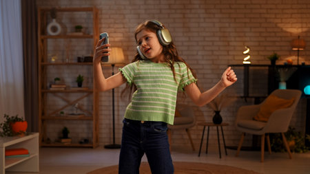 Young girl child spending evening time at home, girl stands in living room listening music in headphones watching video on smartphone and dancing.の写真素材
