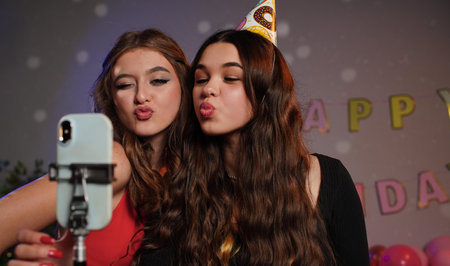 Teenagers birthday party, girls in festive hats posing at camera taking selfie photos and videos,...