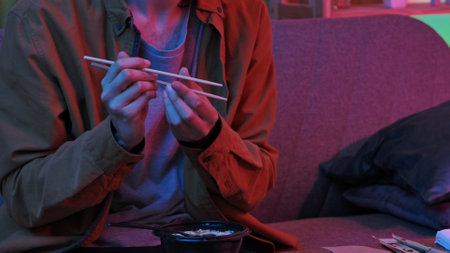 Young man sitting at home in evening on the sofa, ordered delivery of tasty asian food, holding...