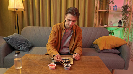 Young man sitting at home in evening on the sofa, ordered delivery of tasty asian food, eating sushi rolls with chopsticks, dipping in soy sauce.の写真素材