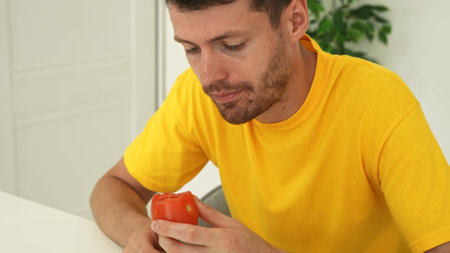 Young man sitting at the table at home and eating fresh vitamin veggies, eating ripe red tomato, chewing and looking, enjoying snack, close shot.の写真素材