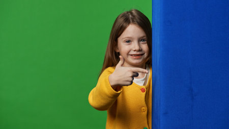 Smiling girl child peeks out and pointing at blue space, positive expression, isolated on chroma key green screen studio background mockup.の写真素材