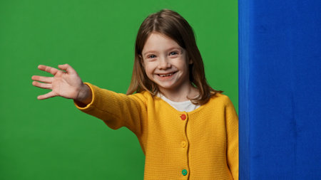 Smiling girl child peeks out and waves hand, positive expression, isolated on chroma key green screen studio background mockup.の写真素材
