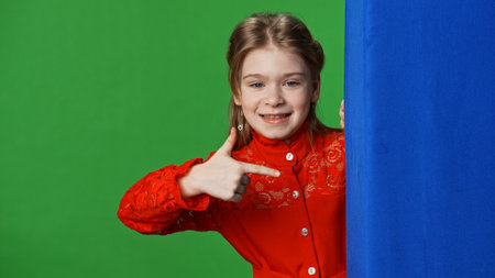 Smiling girl child peeks out and pointing at blue space, positive expression, isolated on chroma key green screen studio background mockup.の写真素材