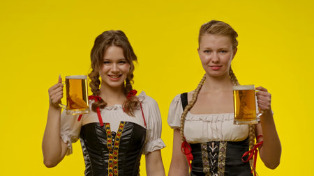 Two Women in Traditional Dresses Holding Beer Mugs in Front of Yellow Backgroundの写真素材