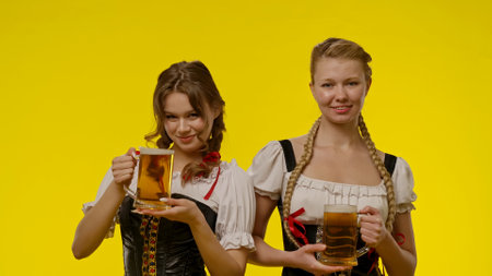 Two Women in Traditional Dresses Holding Beer Mugs in Front of Yellow Backgroundの写真素材