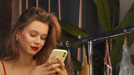 Young Woman Browsing Smartphone Among Hanging Dressesの写真素材