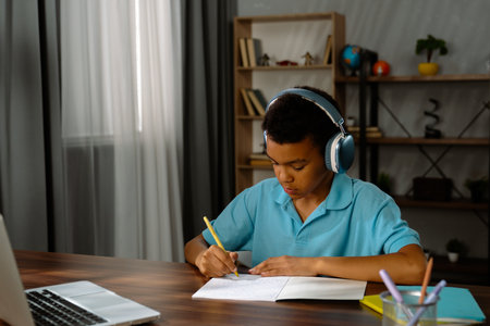 Young african american child boy studying at home, kid in headphones sits at desk attends school...