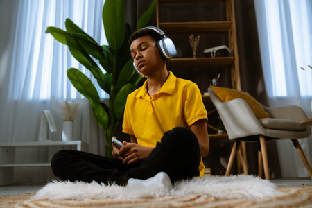 Young african american child boy spending free time at home, kid sits on the floor in the living room and listening music in headphones on smartphone.の写真素材