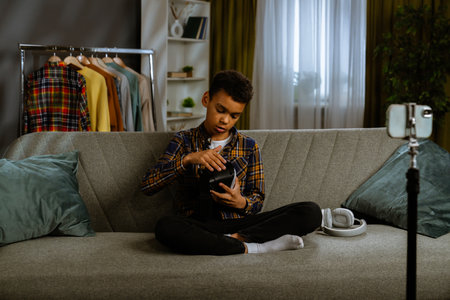 Young african american child boy spending time at home, kid blogger sits on sofa records video on smartphone, talking at camera shows paper sheet with like and subscribe text.の写真素材