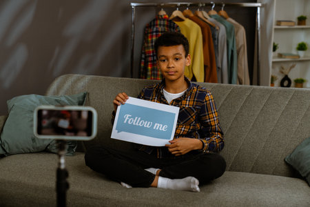 Young african american child boy spending time at home, kid blogger sits on sofa records video on smartphone, talking at camera shows paper sheet with follow me text.の写真素材