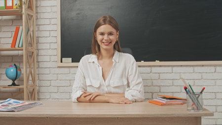 Young woman college teacher sitting at desk in classroom in front of chalkboard, looks at camera smiling, positive expression. Education concept.の写真素材
