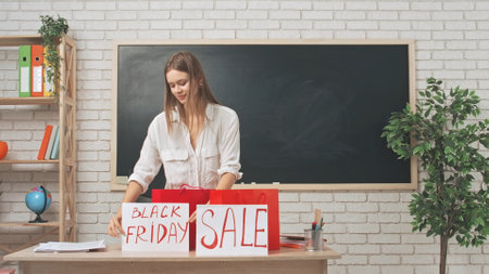 Young woman college teacher walks in standing at desk in classroom in front of chalkboard with gift bags, shows paper sings with sale and black friday written on it.の写真素材