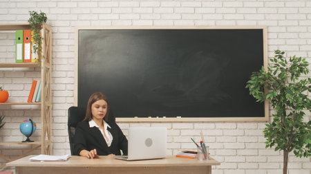 Young woman college teacher at desk in classroom in front of chalkboard explains lesson to students...