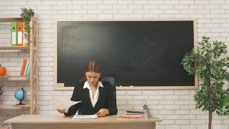 Young woman college teacher at desk in classroom in front of chalkboard reading grading homework or tests of students. Education concept.の写真素材