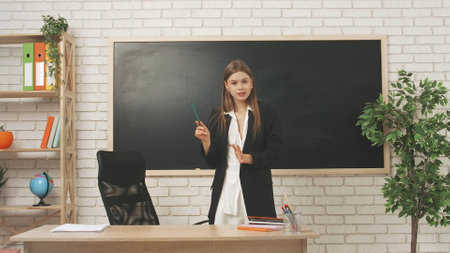 Young woman college teacher standing at desk in classroom in front of chalkboard talking and explains lesson to students. Education concept.の写真素材